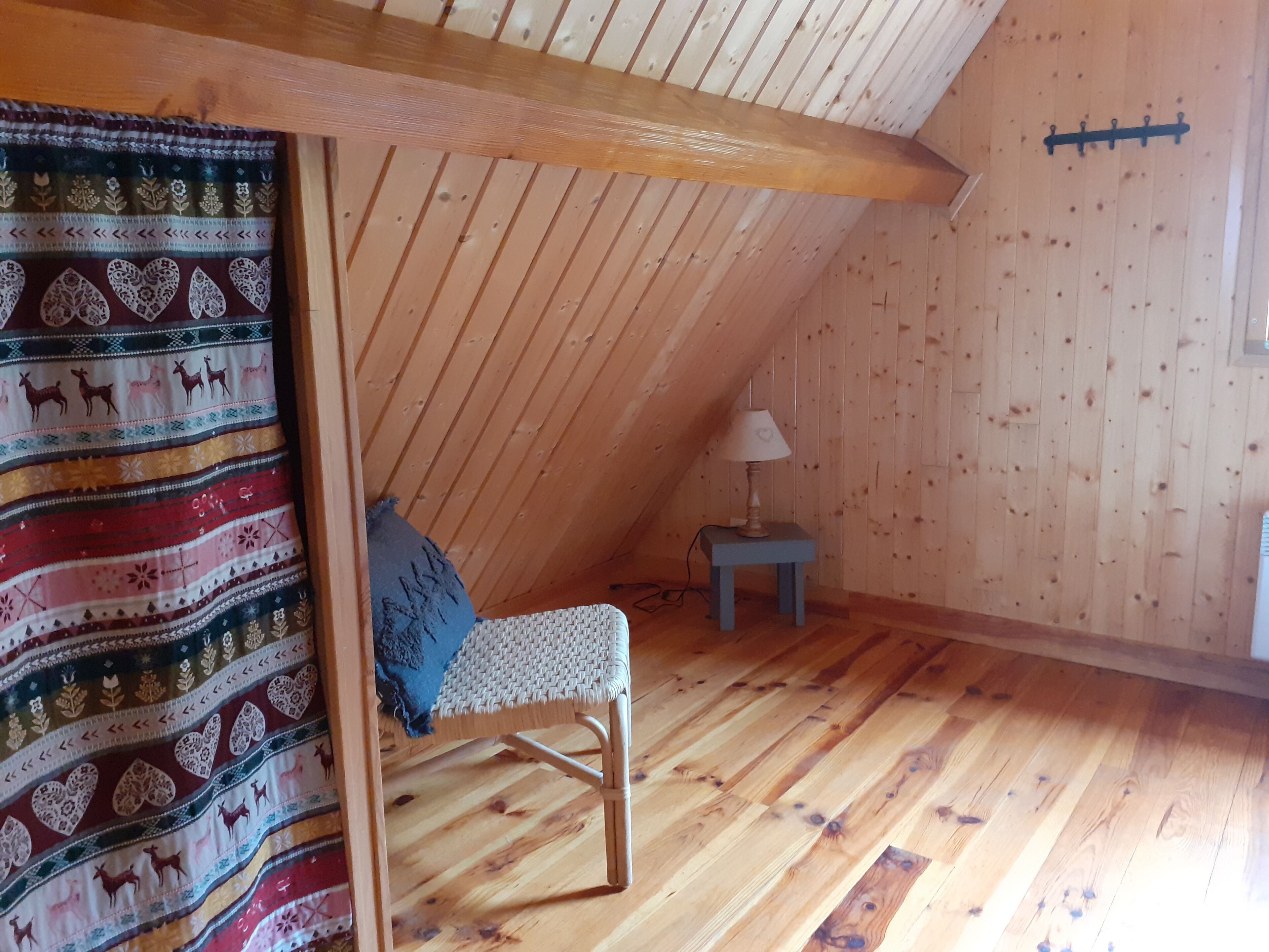 A cozy attic room with wooden walls and a sloped ceiling. A woven chair with a blue cushion is positioned against the wall, and a small table with a lamp is nearby.