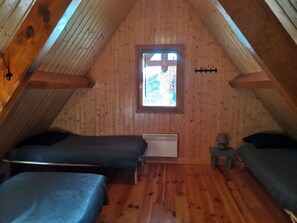 Une petite chambre mansardée, chaleureuse et accueillante, avec des murs en bois et un plafond en pente. On y trouve deux lits simples avec une literie sombre, un petit radiateur et une fenêtre équipée de stores.