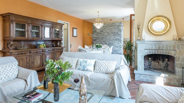 A living room with a sofa set, a wooden cabinet, a dining area with a table set for four, and a fireplace. - Casa Maravilla 6 in Castelnuovo Berardenga (Castelnuovo Berardenga)