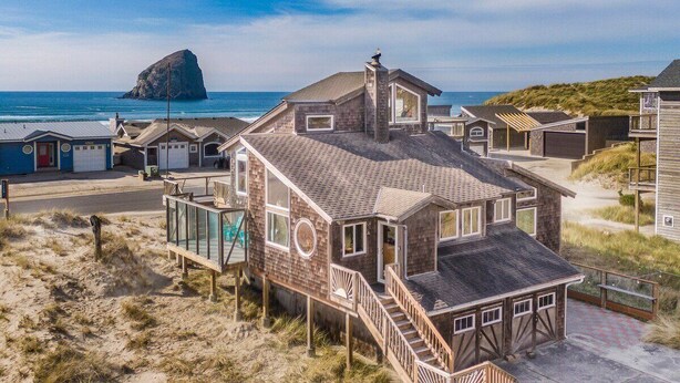 Una casa sulla spiaggia con un'ampia veranda, diversi portoni di garage e una grande formazione rocciosa sullo sfondo.