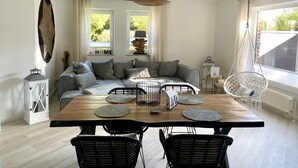 Un salon moderne avec un canapé, une table à manger et des chaises. Il y a deux fenêtres avec des rideaux, un luminaire suspendu et un radiateur.