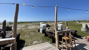 Un espace extérieur aménagé avec des tables et des chaises en bois, des guirlandes lumineuses et une vue sur la plage et le ciel.