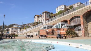 A coastal promenade with picnic tables, a swimming pool, and a stone bridge.