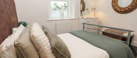 A bedroom with a bed, pillows, a dresser, a mirror, and a window with plants.