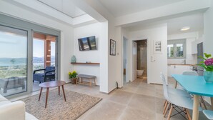A modern living room with a large sliding glass door leading to a balcony, a dining area with a blue table, and a kitchen visible through an open door. - 31 Blue - Aqua Blue - Private Infinity Pool and BBQ Area (Apolpena)