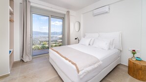 A bedroom with a large bed, a view through a sliding door, a round mirror, and a wooden side table with a vase. - 31 Blue - Aqua Blue - Private Infinity Pool and BBQ Area (Apolpena)