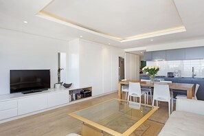 A modern living room with a flat-screen TV, a glass-top dining table, white chairs, a sofa, and a kitchen area with a sink and cabinets. - Nine On S 3E (Cape Town)