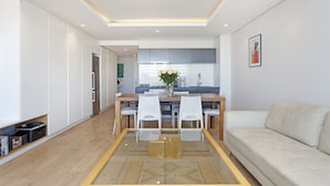 A modern living room with a glass dining table, white chairs, a beige sofa, and a kitchen in the background. - Nine On S 3E (Cape Town)