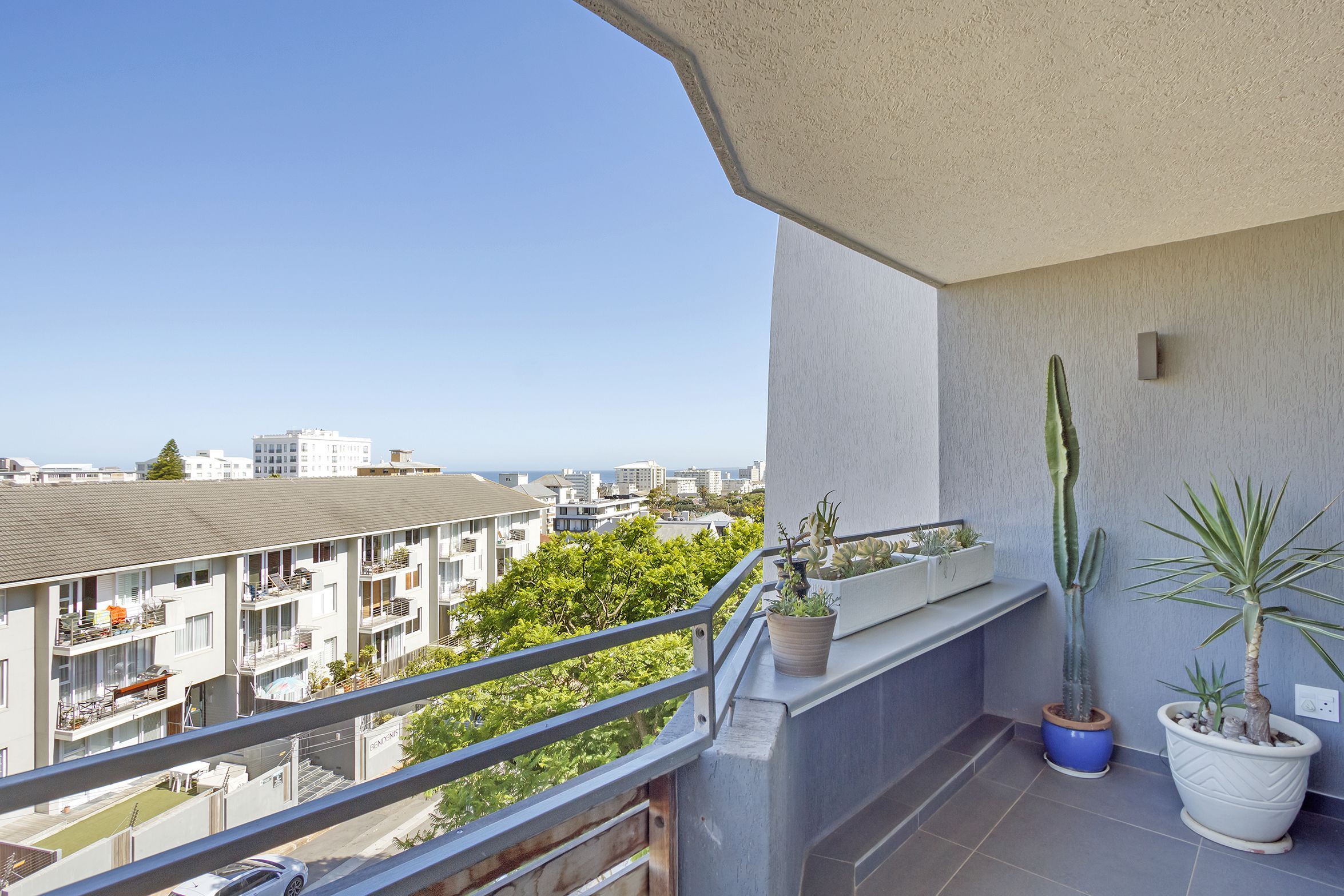 A balcony with potted plants, a railing, and a view of buildings and greenery.
