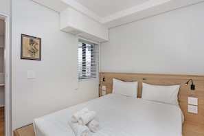 A neatly made bed with white linens and rolled towels, a wooden headboard, and a wall-mounted light fixture.