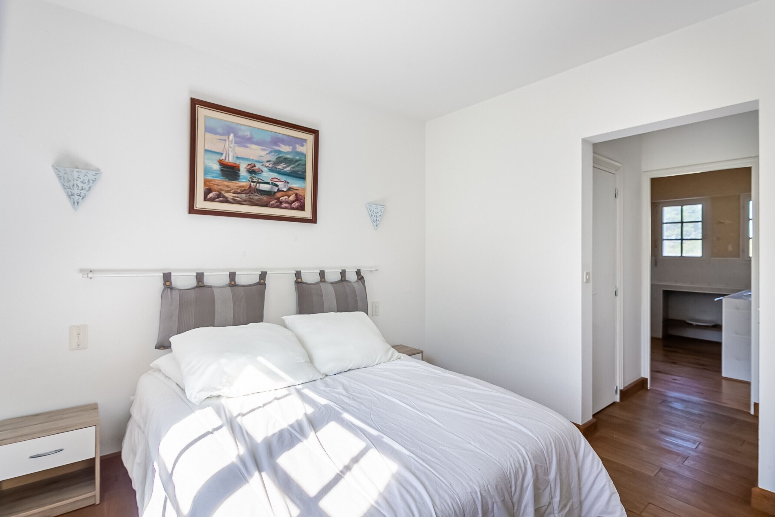 A bedroom with a bed, a nightstand, a drawer, a painting, and a hallway leading to another room.