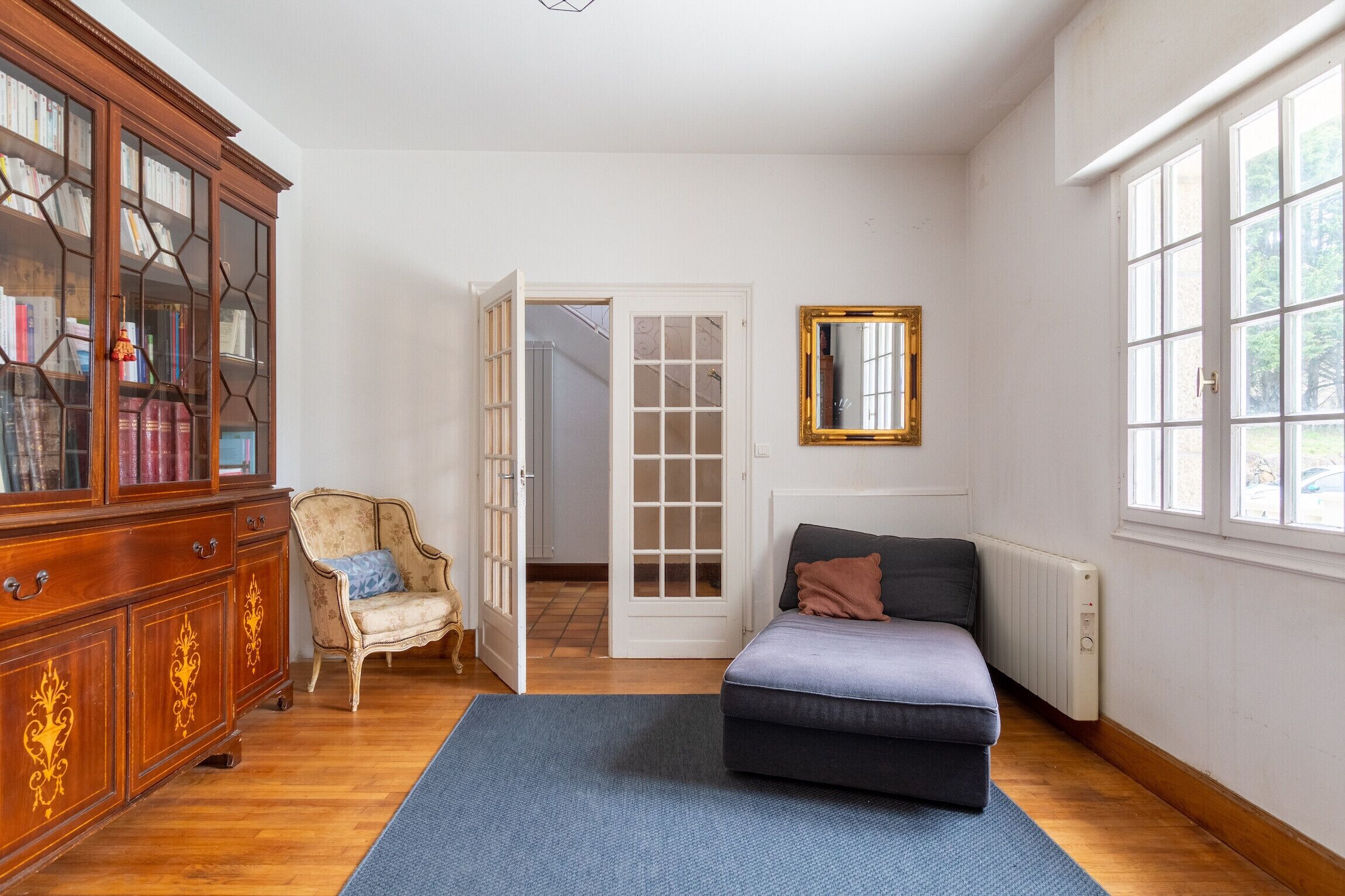 Una habitación con un armario de madera, una silla, un sofá, un espejo y una puerta que da a otra estancia.