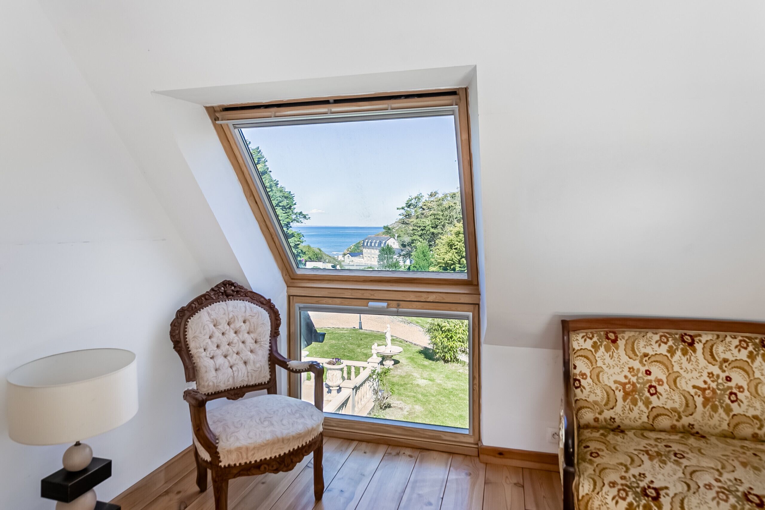 A room with a sloped ceiling, a large window offering a view of the sea, a wooden floor, a tufted chair, and a patterned sofa.