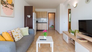 Ein modernes Wohnzimmer mit einer grauen Couch, einem weißen Couchtisch mit Blumenvase, einem Fernseher auf einem Holzständer und einer Küche im Hintergrund.