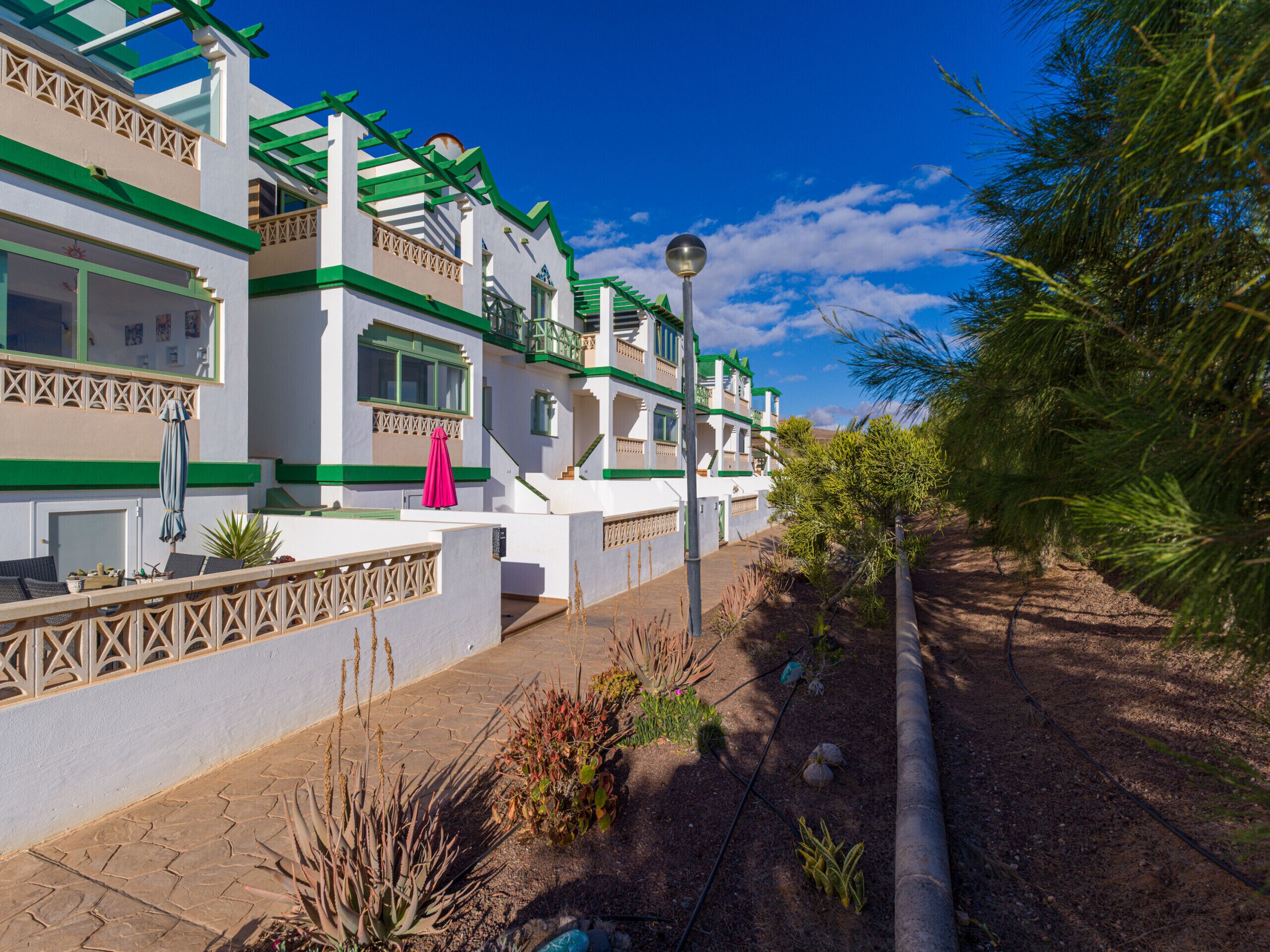 A row of white buildings with green accents and balconies, a paved walkway, and some landscaping.