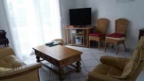 A living room with a wooden coffee table, two yellow leather sofas, a television on a stand, and wicker chairs with red cushions. - 4-bedroom house in the heart of the Abers (7-minute walk from Ganaoc beach) (Saint-Pabu)
