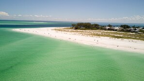 An aerial view of a beach with clear turquoise waters, a sandy shore, and a cluster of buildings in the background. - Bayview Inn #101 · Heated Pool · Bridge St (Bradenton Beach)