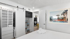 Ein Hotelzimmer mit einem Bett, einem weißen Kleiderschrank, einem Schreibtisch und einem Gemälde einer Strandansicht.