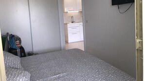 Een kleine, smalle kamer met een bed, een aan de muur bevestigde televisie en uitzicht op een keuken met witte kasten en een spoelbak.