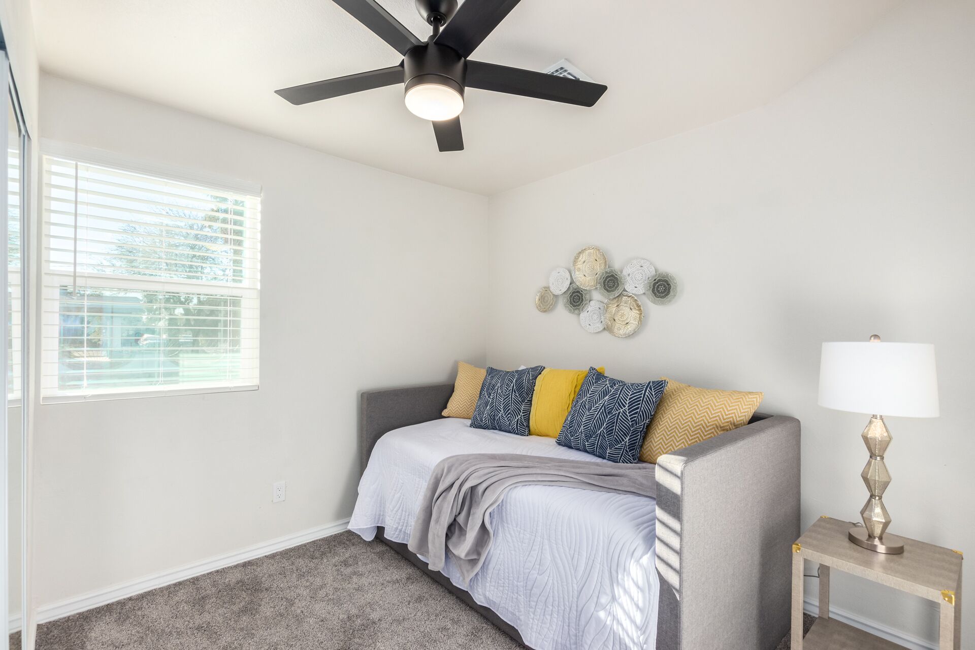 Une chambre à coucher comprenant un lit, un ventilateur de plafond, une fenêtre avec des stores, un objet décoratif fixé au mur et une table de chevet avec une lampe.
