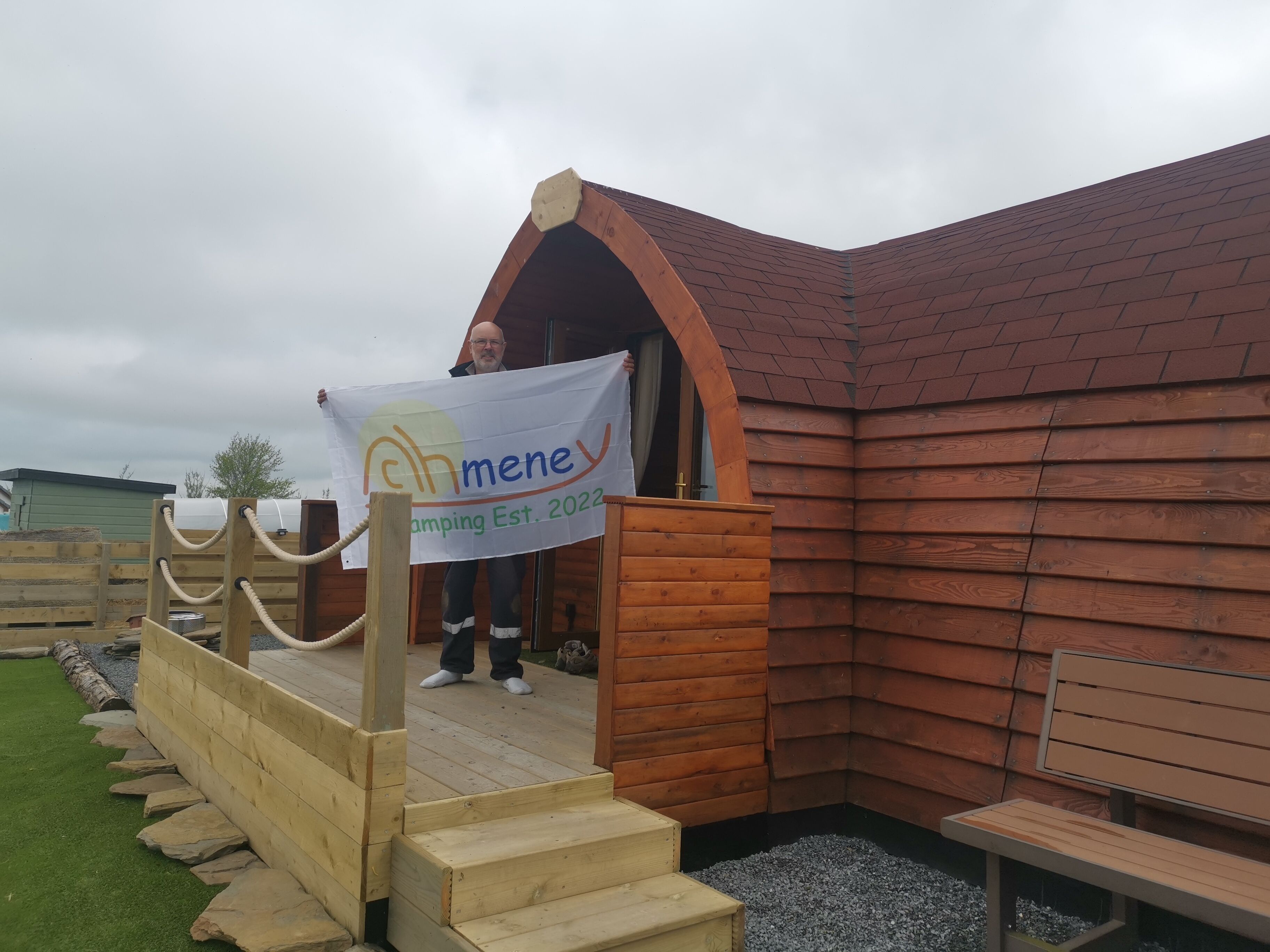 A wooden cabin with a ramp leading to a porch, a bench, and a banner indicating the establishment year 2022.