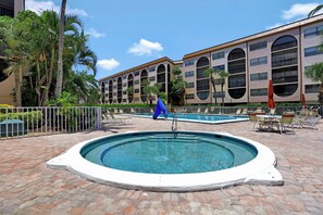 A swimming pool with a blue chair and a person sitting on a ladder. There are palm trees and a building with large windows in the background. - Waterfront Condo in Anglers Cove | Pools & Spas (Marco Island)