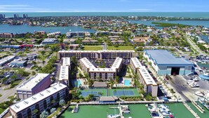 Vue aérienne d’une ville côtière, où l’on aperçoit un hôtel imposant doté d’une cour centrale, de courts de tennis et d’un port de plaisance avec des bateaux.