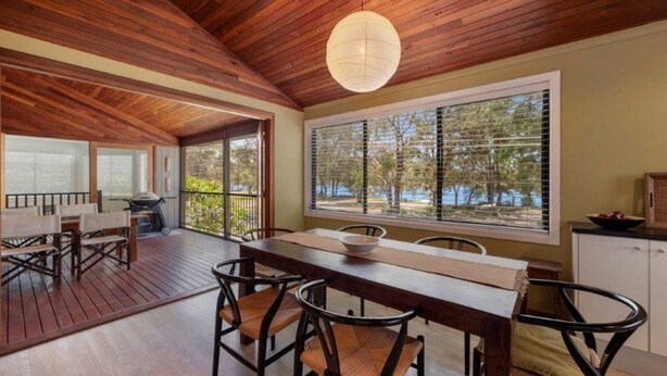 A wooden deck with a dining table and chairs, a grill, and a large window with blinds. - Lakeside Lodge - Fabulous Waterfront Holiday (Laurieton)