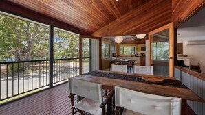 A wooden deck with a dining table and chairs, a kitchen area, and large windows offering a view of trees and a road. - Lakeside Lodge - Fabulous Waterfront Holiday (Laurieton)