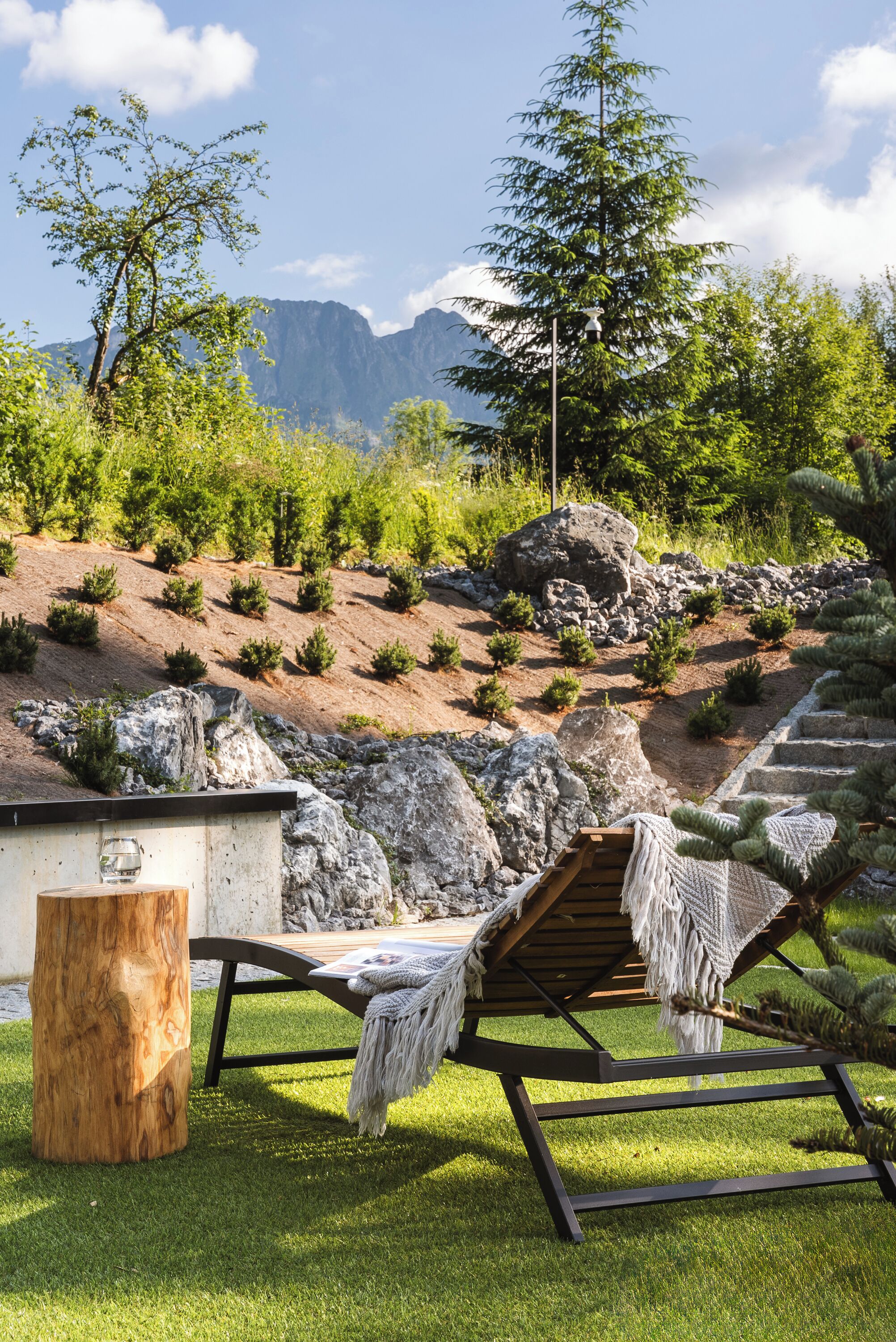 A lounge chair with a blanket, a wooden table, and a stone stump.