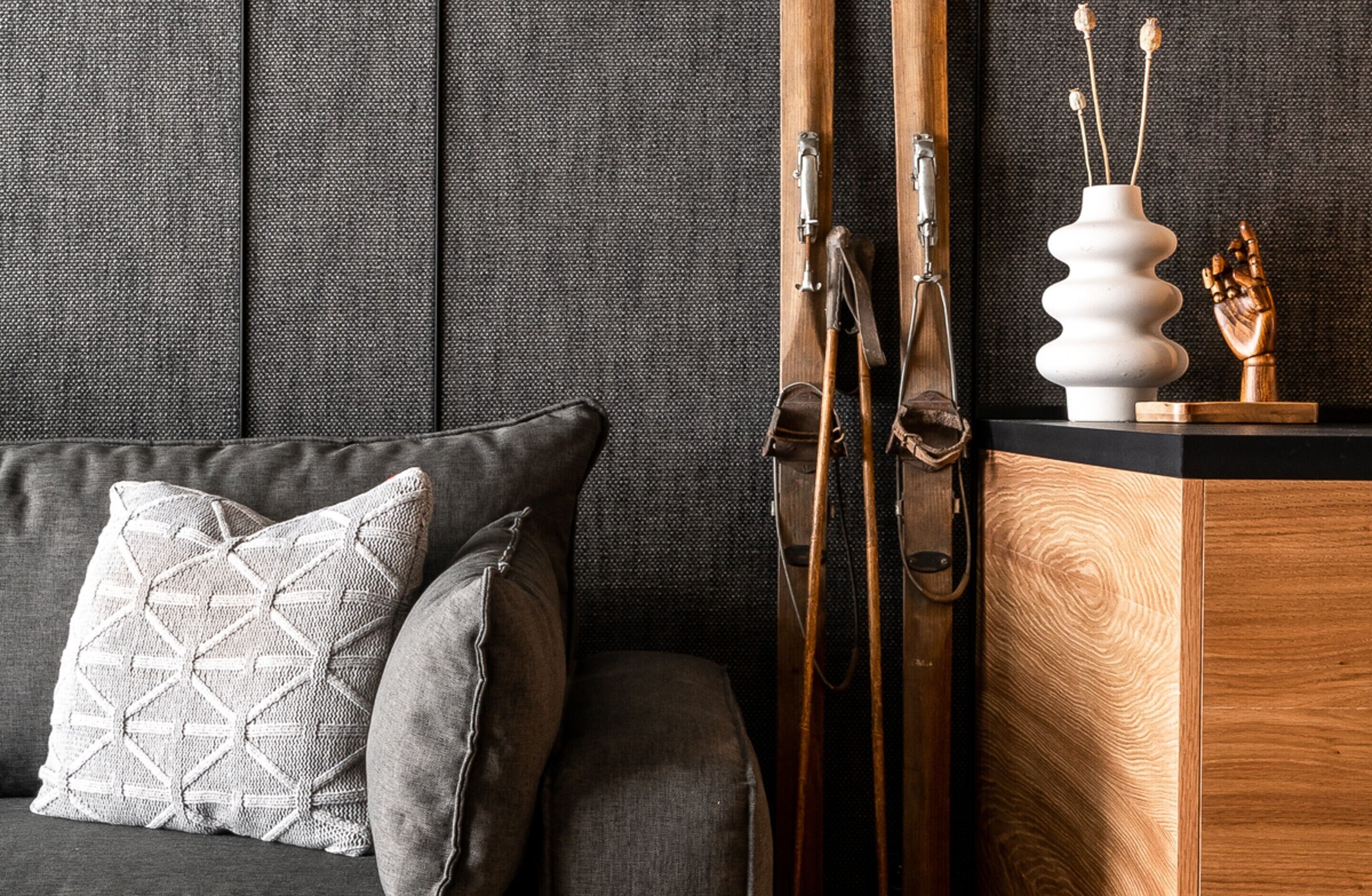 A room with a grey sofa, a patterned pillow, a wooden cabinet, and a pair of skis leaning against the wall.