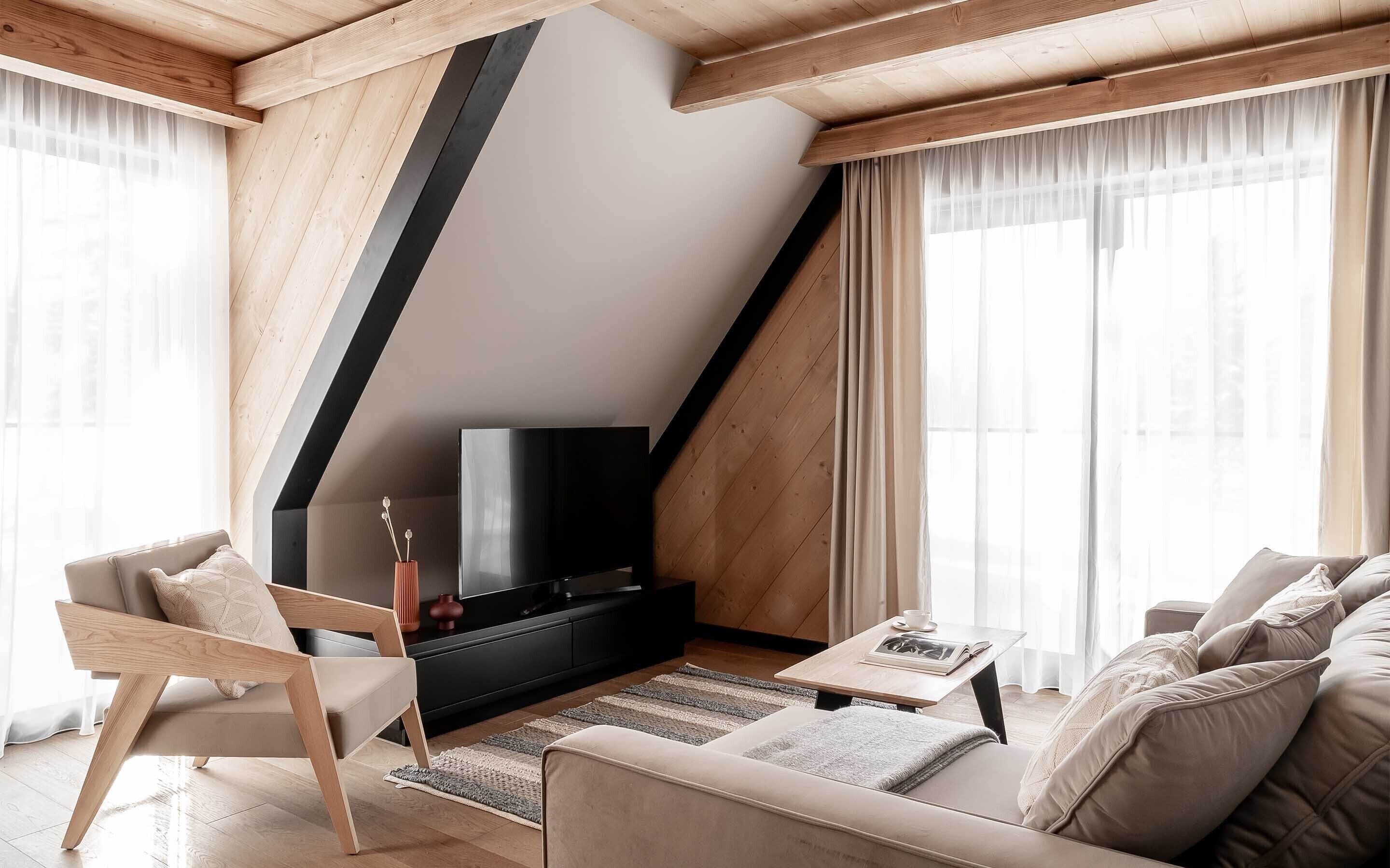Un salón moderno con techo inclinado, vigas de madera, mueble de entretenimiento negro, sofá color beige y una mesa de centro.