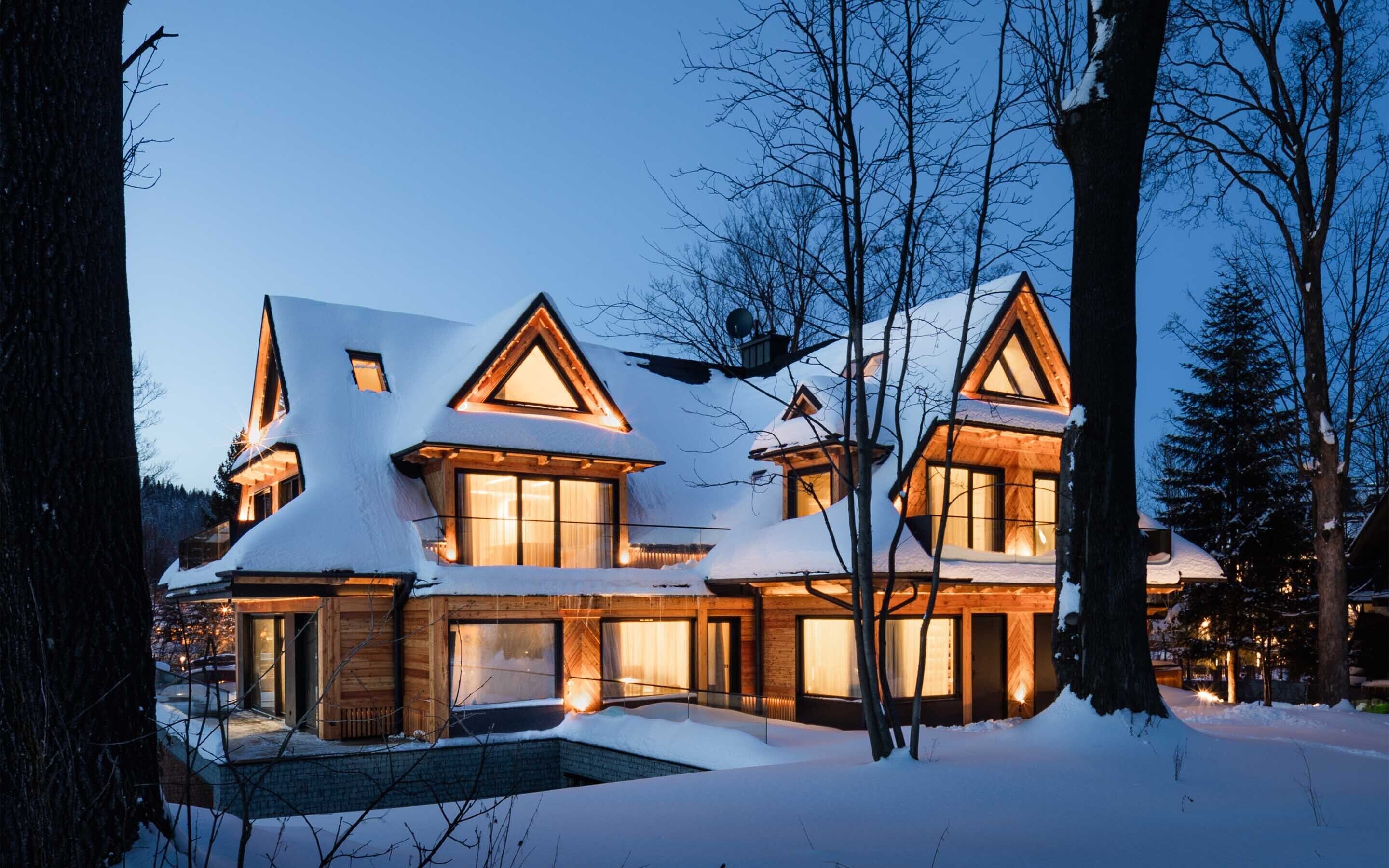 A large, multi-story wooden house with a snow-covered roof and large windows, surrounded by trees.
