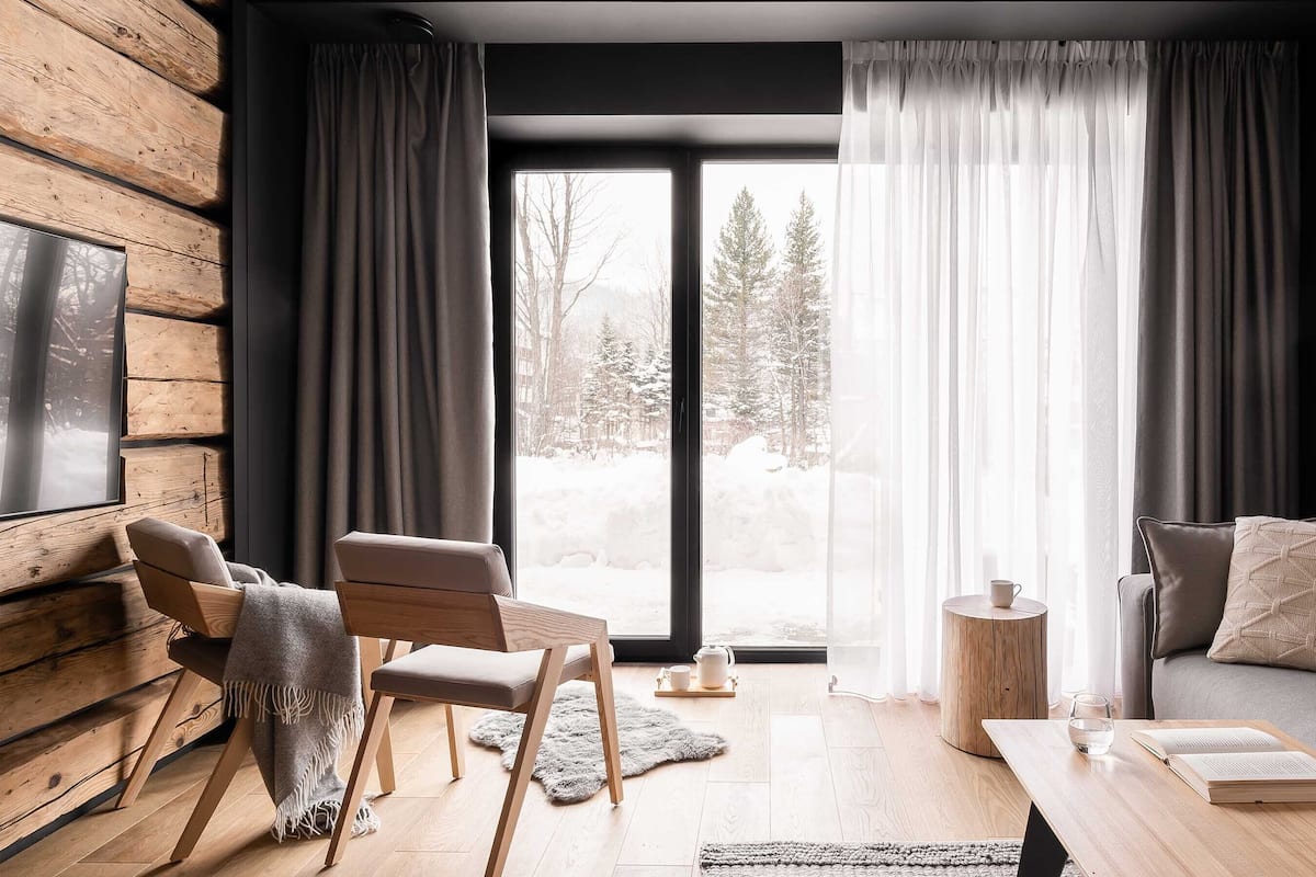 A cozy living room with a wooden wall, a sofa, a chair, a coffee table, and a view of a snowy landscape through large windows.