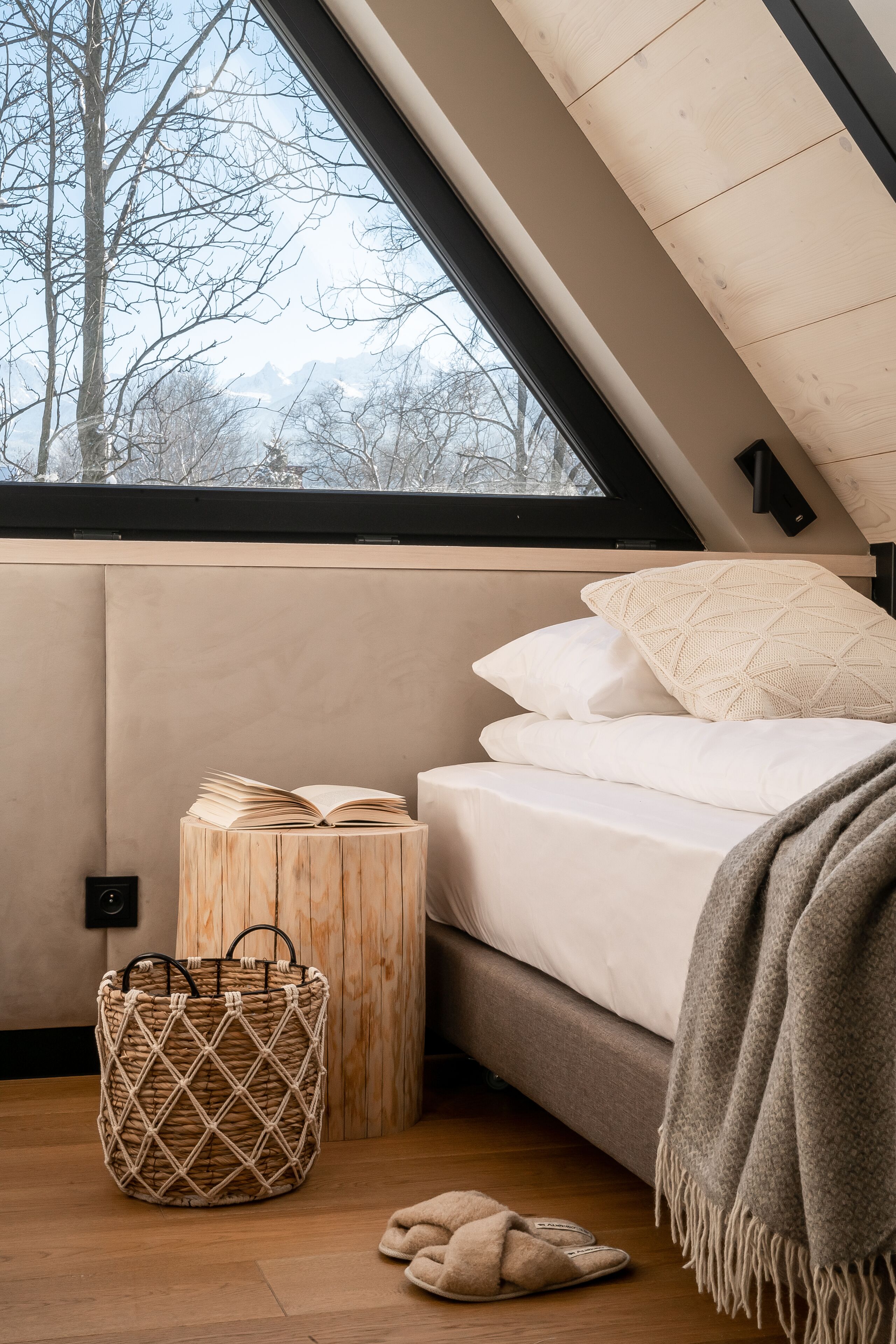 A bedroom with a sloped ceiling, a bed with white bedding, a woven basket, and a view of trees and mountains through a window.