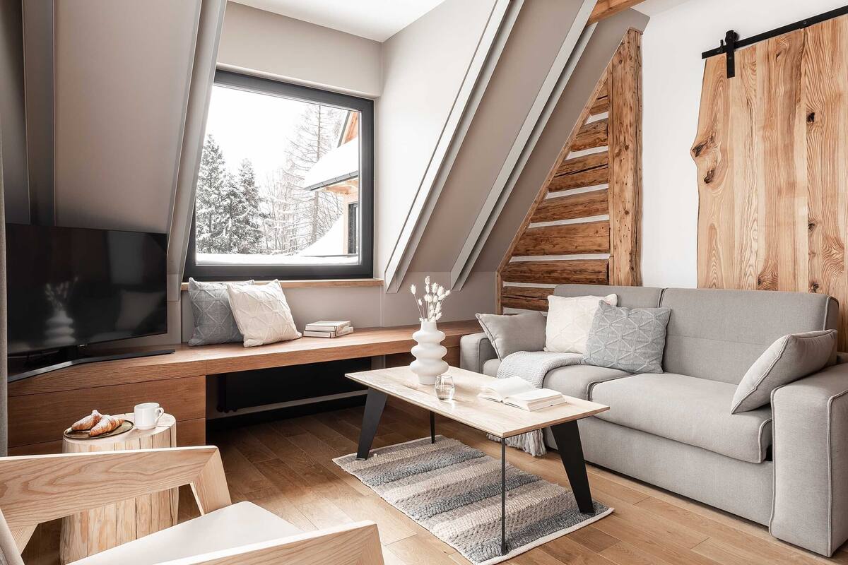 A modern loft-style living room with a large window, a grey sofa, a wooden coffee table, and a TV on a wooden stand.