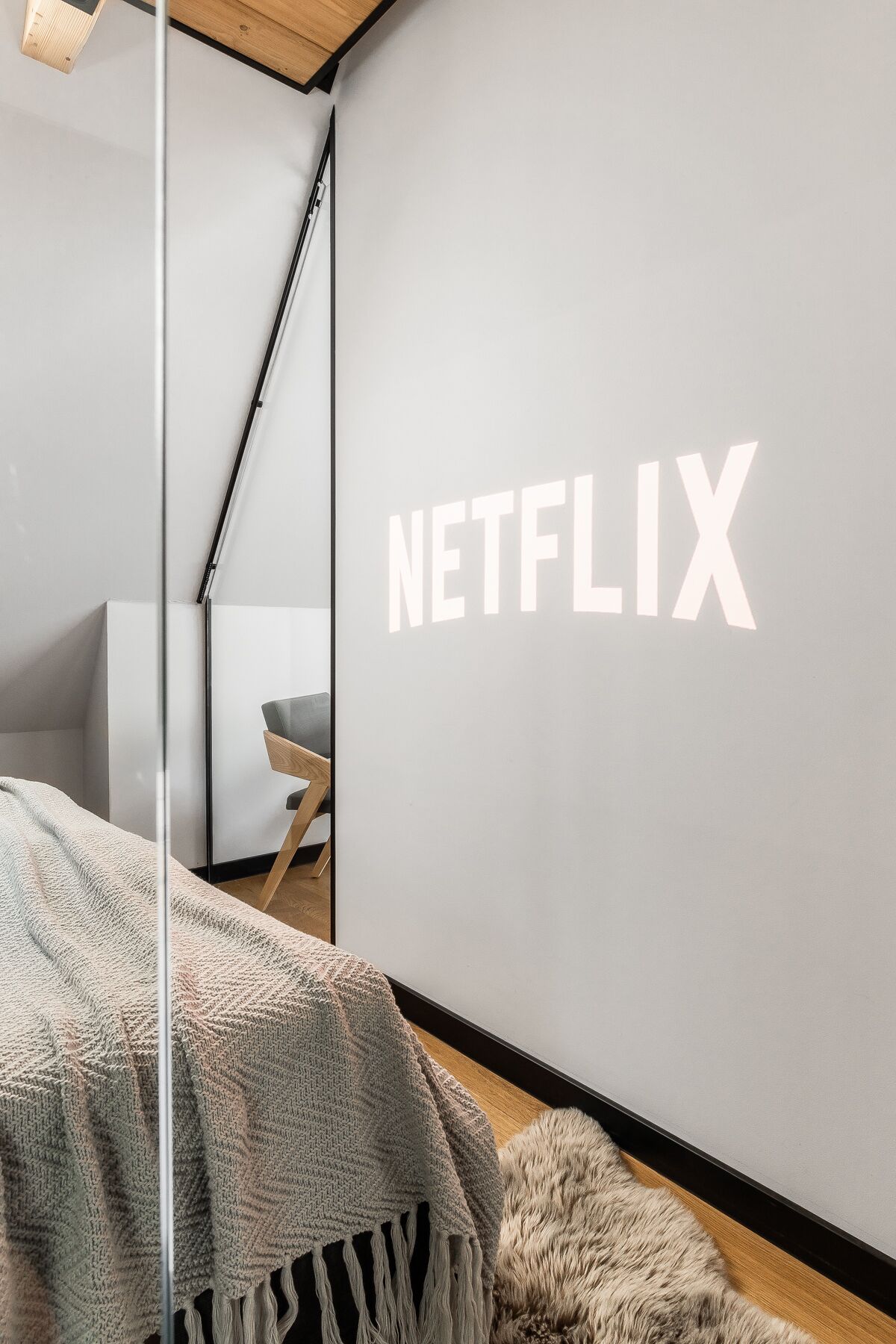 A modern bedroom with a large wall-mounted screen displaying "NETFLIX," a cozy bed with a knitted blanket, a wooden floor, and a minimalist chair.
