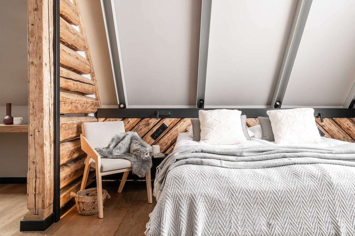 A cozy attic bedroom with a sloped ceiling, wooden beams, a comfortable chair, a bed with a white blanket, and a small table with a cup and vase.