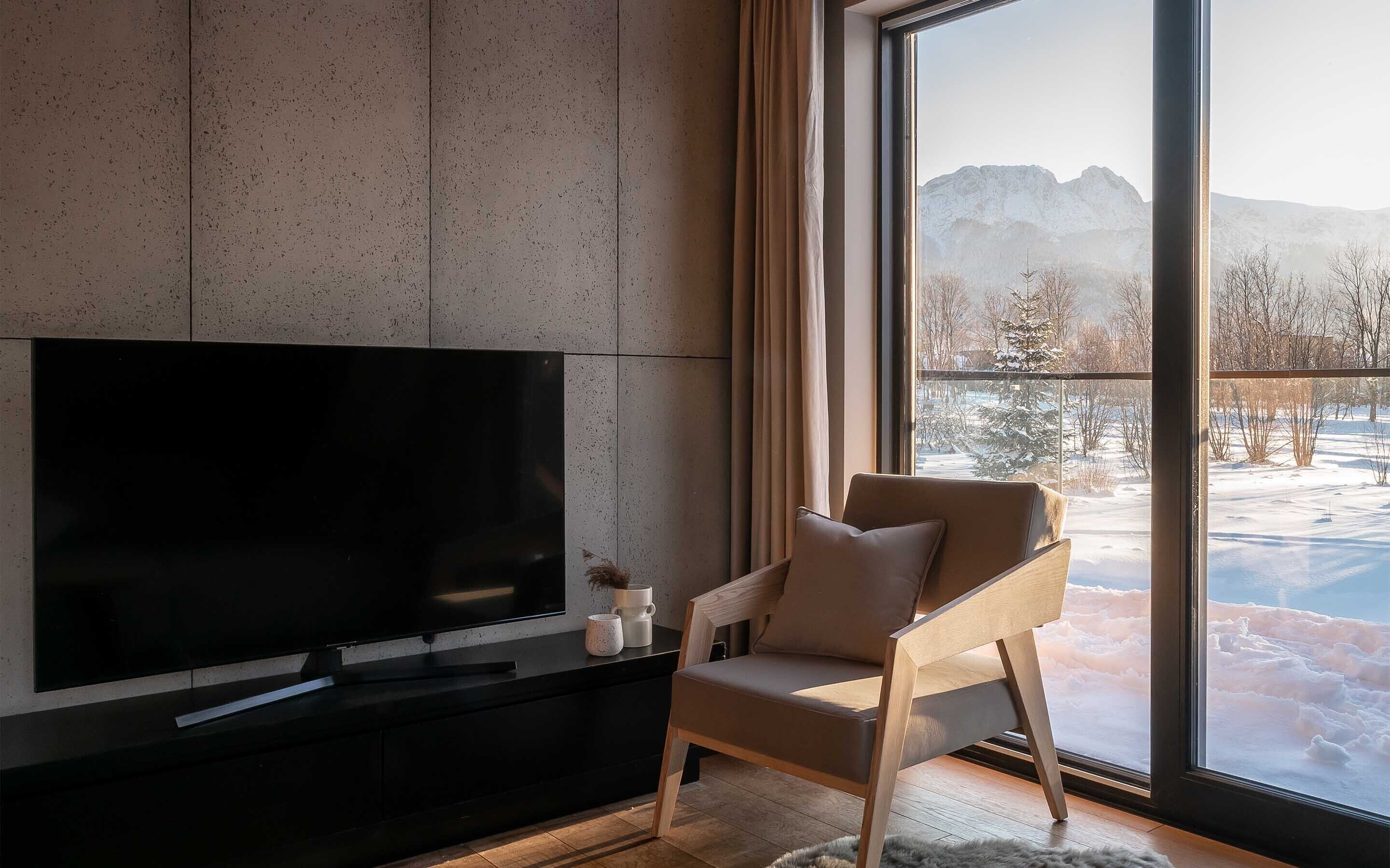 Un salon moderne avec un grand téléviseur à écran plat posé sur un meuble noir, un fauteuil beige et une grande fenêtre donnant sur un paysage de montagne enneigé.