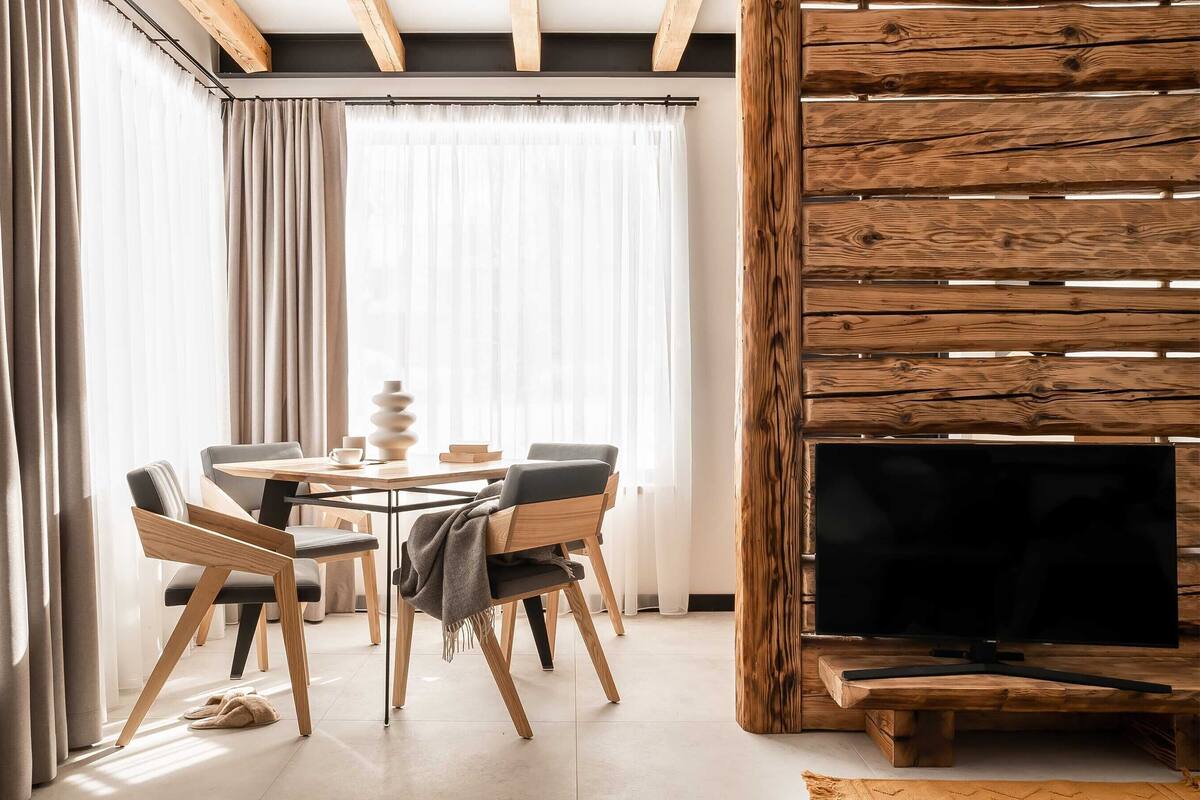 A modern living room with a wooden TV stand, a dining area with a round table and chairs, and a large window with sheer curtains.