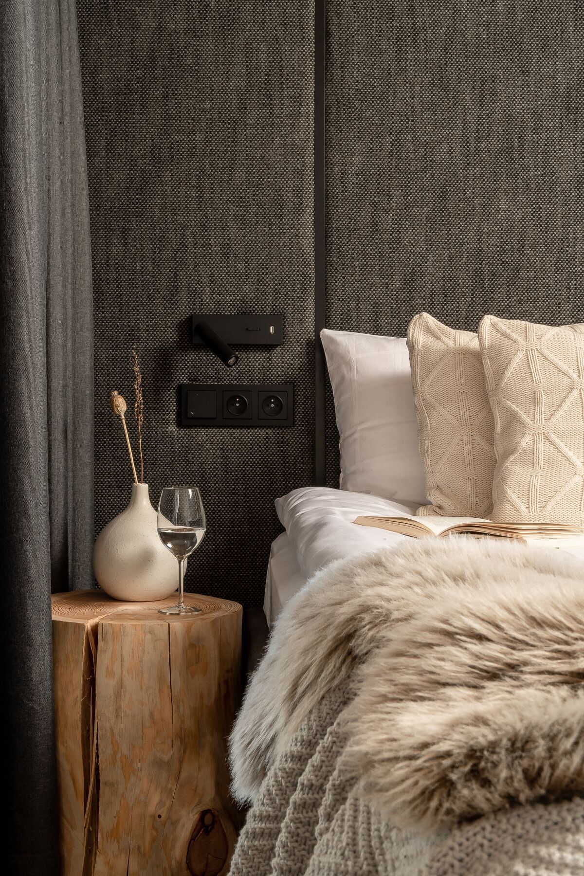 Un dormitorio con una mesita de noche de madera, un florero con ramas secas, una copa de vino y una cama con una colcha blanca y un cojín de punto.