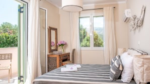 A bedroom with a bed, a desk, a chair, a window with curtains, and a view of greenery outside. - Villa Athena (Corfu)