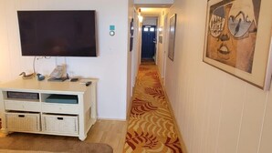 A hallway with a patterned carpet, a flat-screen TV, a white console table, and framed artwork on the walls. - Calypso unit 602, 6201 Coastal Hwy., Ocean City, Maryland 21842 (Ocean City)