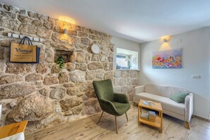 A room with a stone wall, a green chair, a white sofa, a wooden side table with books, and a painting on the wall.