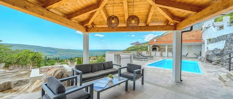 A covered outdoor seating area with a pool, stone flooring, and a view of the landscape.