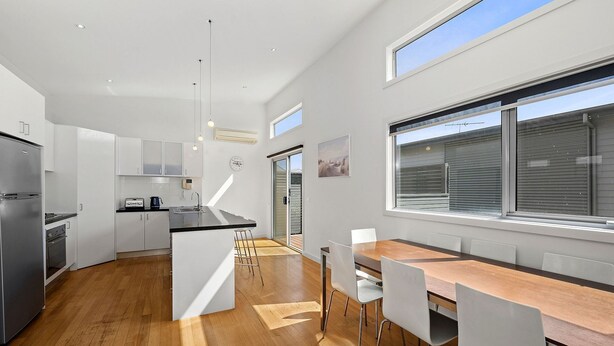 A modern kitchen with a central island, white cabinetry, and a dining area with a wooden table and chairs. - Anderson Central Townhouse - Walk to Shops & Beach (Torquay)