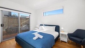 A modern bedroom with a large bed, a side table, a chair, and a window with a view of a fence and trees. - Anderson Central Townhouse - Walk to Shops & Beach (Torquay)