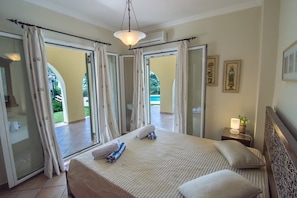 A hotel room with a bed, a nightstand with a lamp, and a view of a pool through open curtains.