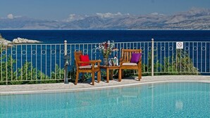 Un espace aménagé au bord de la piscine, avec deux chaises en bois et une table, un vase rempli de fleurs, et une vue sur la mer et les montagnes.