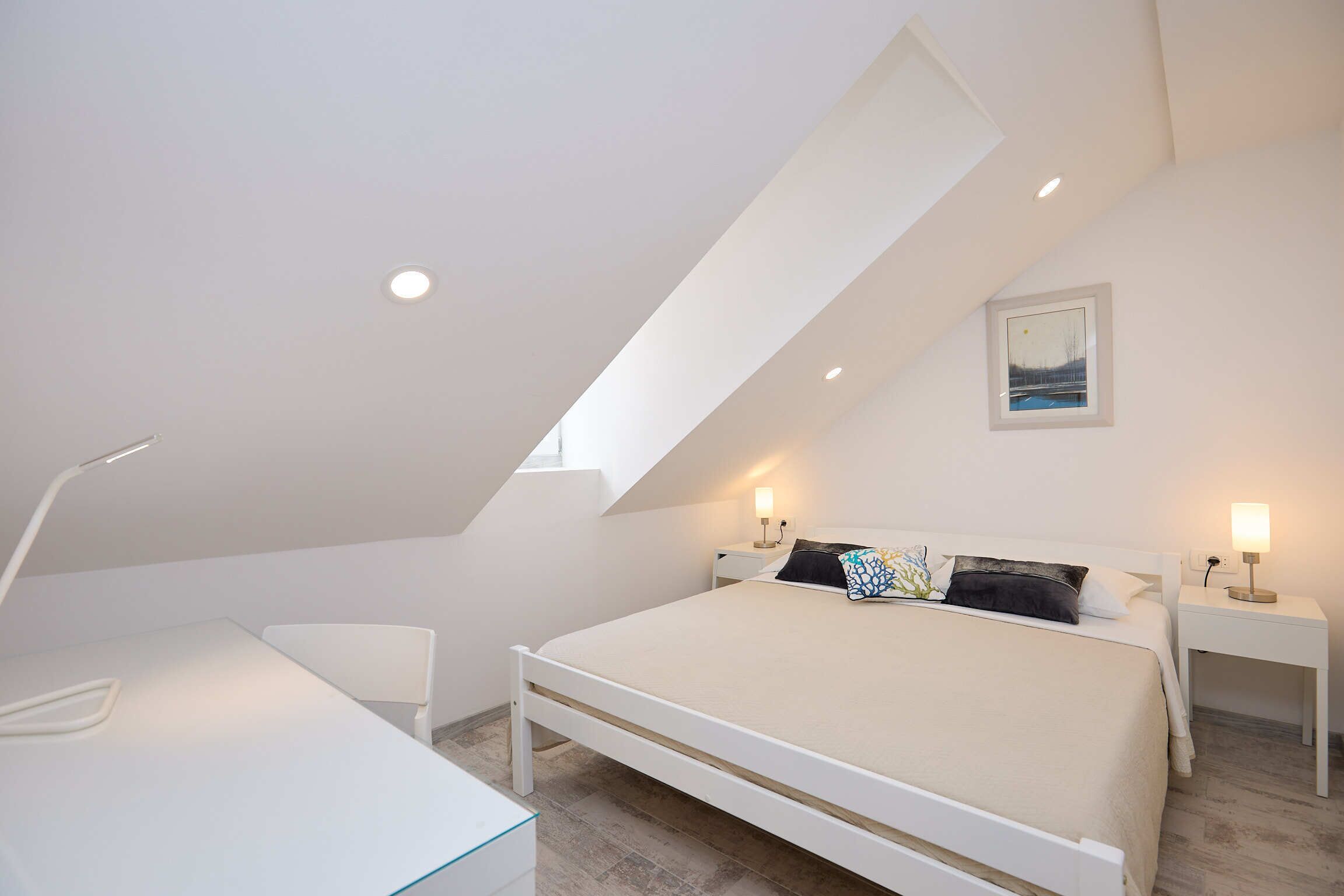A bedroom with a sloped ceiling, a bed, bedside tables with lamps, and a desk.